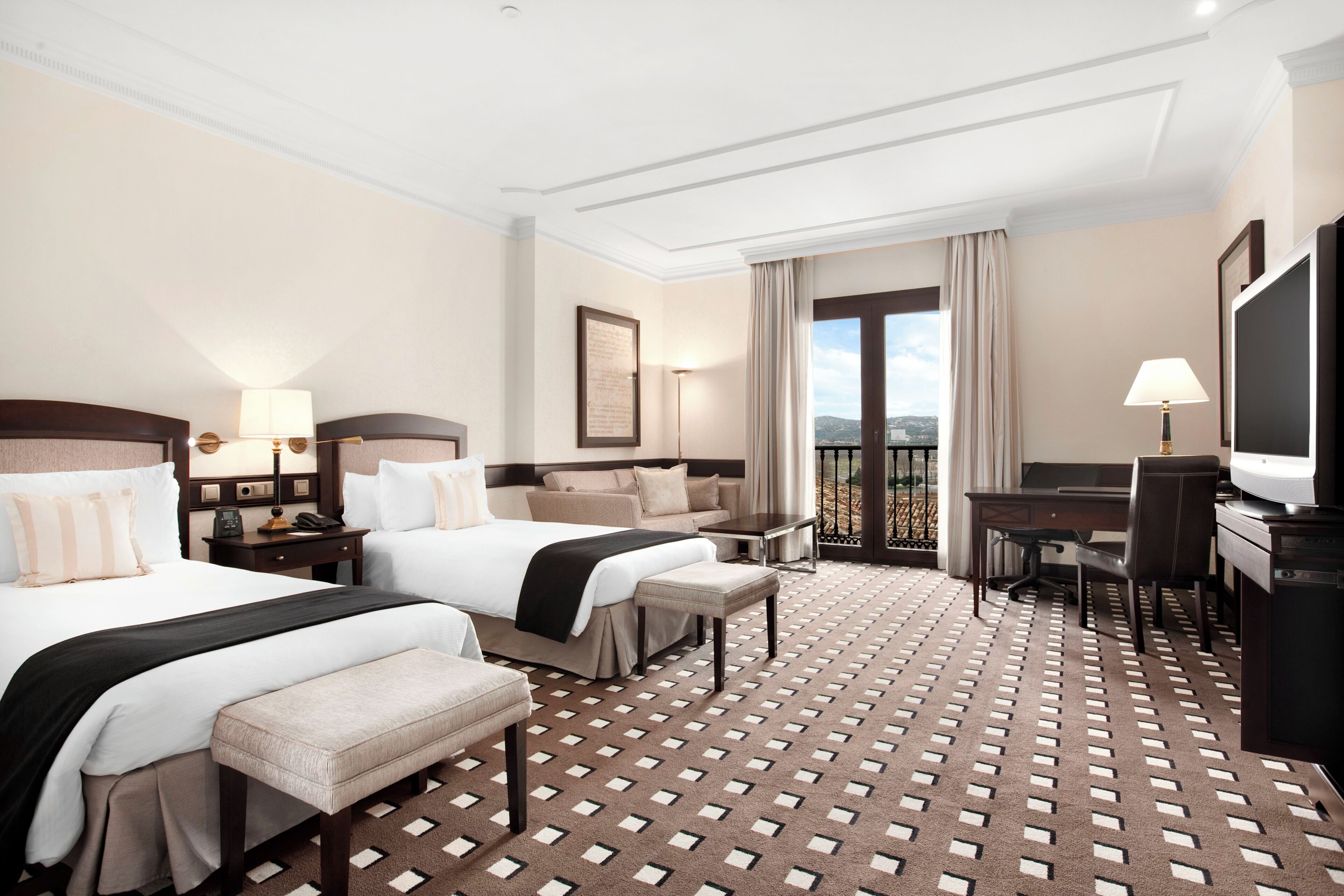 A hotel room with two beds, a desk, a chair, a TV, and a balcony with a view.