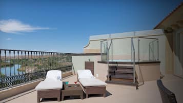 Ein Balkon mit zwei Liegestühlen und einem kleinen Tisch, mit Blick auf ein Gewässer und eine entfernte Landschaft.