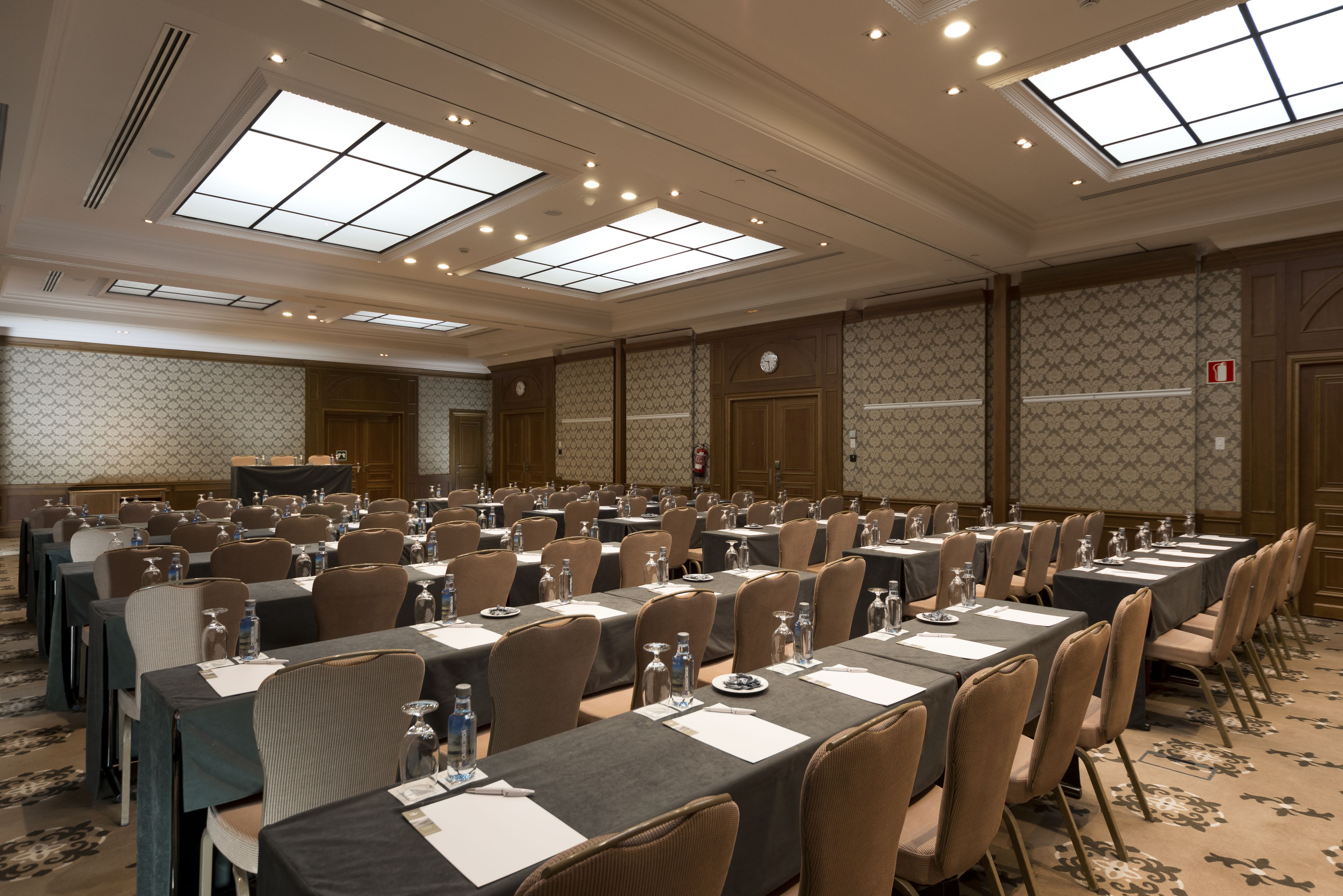 A large conference room with multiple tables and chairs, equipped with water bottles and glasses, and a clock on the wall.