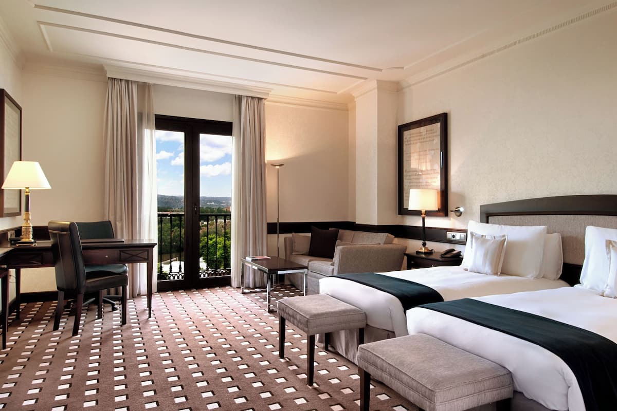 Une chambre d’hôtel équipée d’un bureau, de deux lits, d’une chaise, d’une petite table et d’un balcon avec des rideaux.