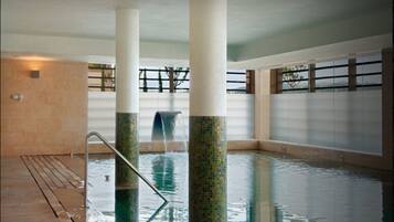 Una piscina interior con suelo de baldosas, columnas con diseños de mosaicos y una amplia vista al exterior a través de grandes ventanales.