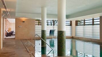 Una piscina interior con una cascada, rodeada de amplios ventanales y suelo de baldosas.
