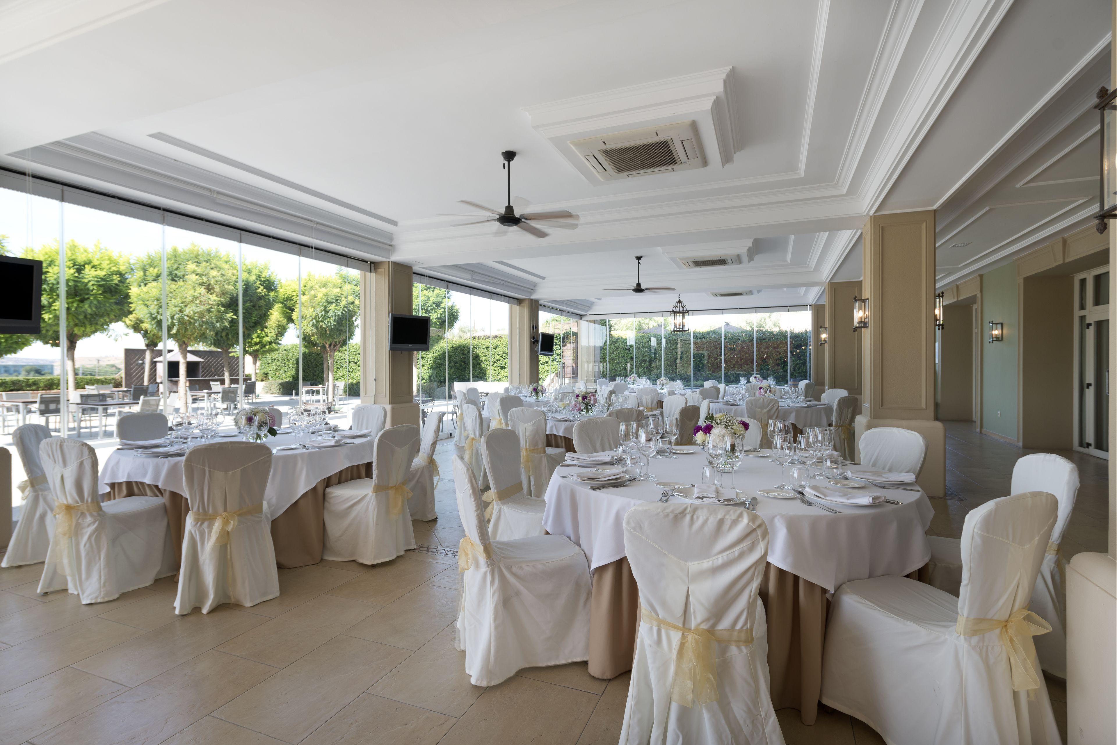 Un salón de banquetes con mesas redondas, sillas blancas con cintas doradas y vistas a zonas verdes al exterior.