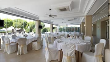 Un salón de banquetes con mesas redondas, sillas blancas con cintas doradas y vistas a zonas verdes al exterior.