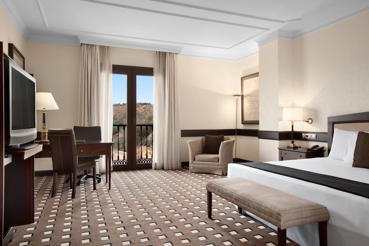 Habitación de hotel con cama, escritorio, silla, televisor y balcón con cortinas.