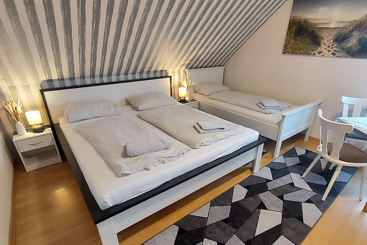 Een moderne slaapkamer met een groot bed, twee kleinere bedden, een bureau en een stoel. Er staan nachtkastjes met lampen en er hangt een schilderij aan de muur.