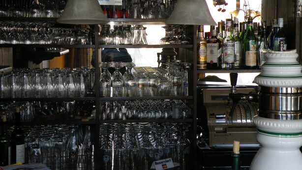 A bar area with shelves of glasses and bottles, a cash register, and signs with German text. - Hotel zum Norden (Jagel)