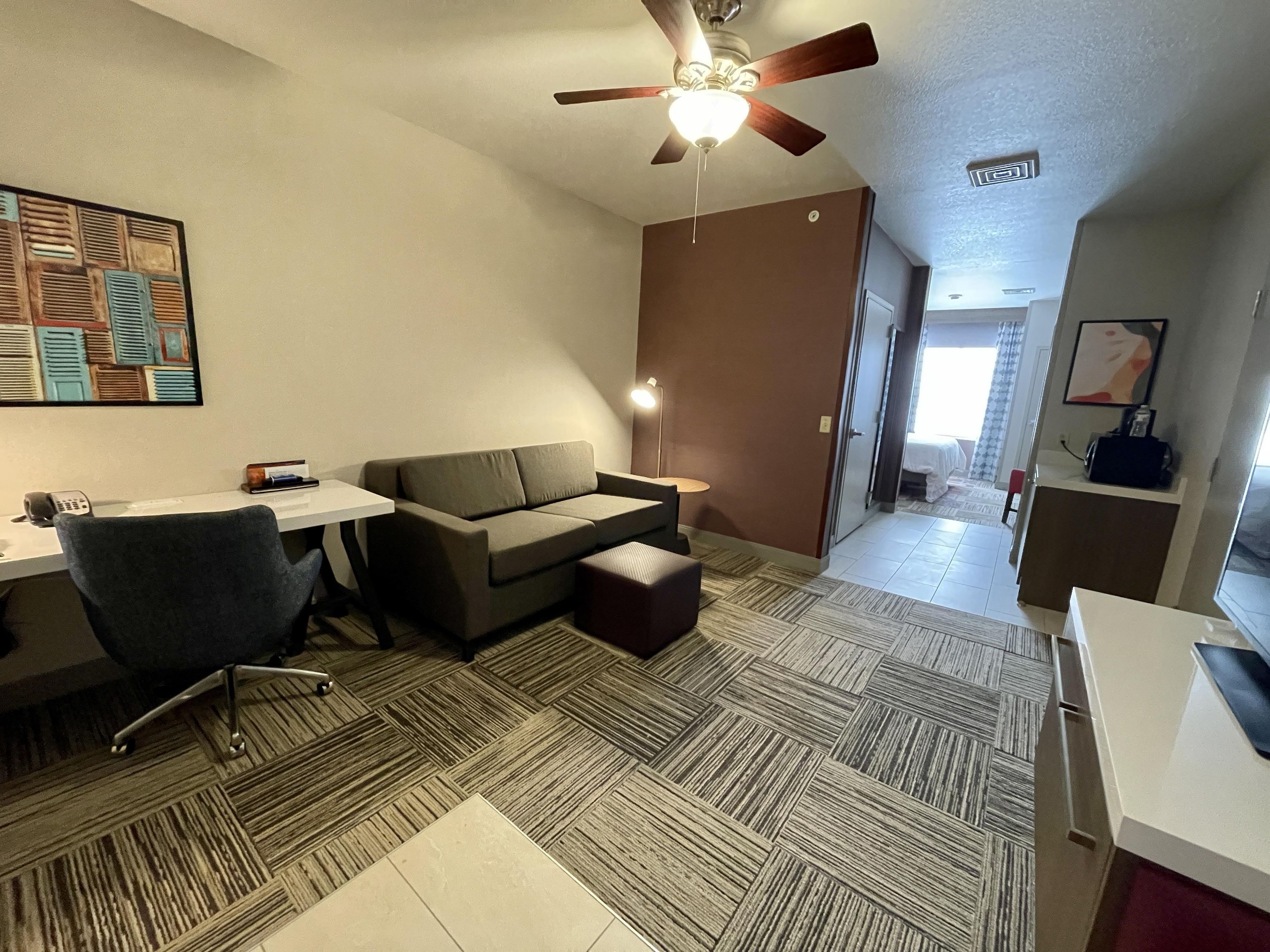 A compact living space with a sofa, a desk, a chair, a ceiling fan, and a patterned floor.