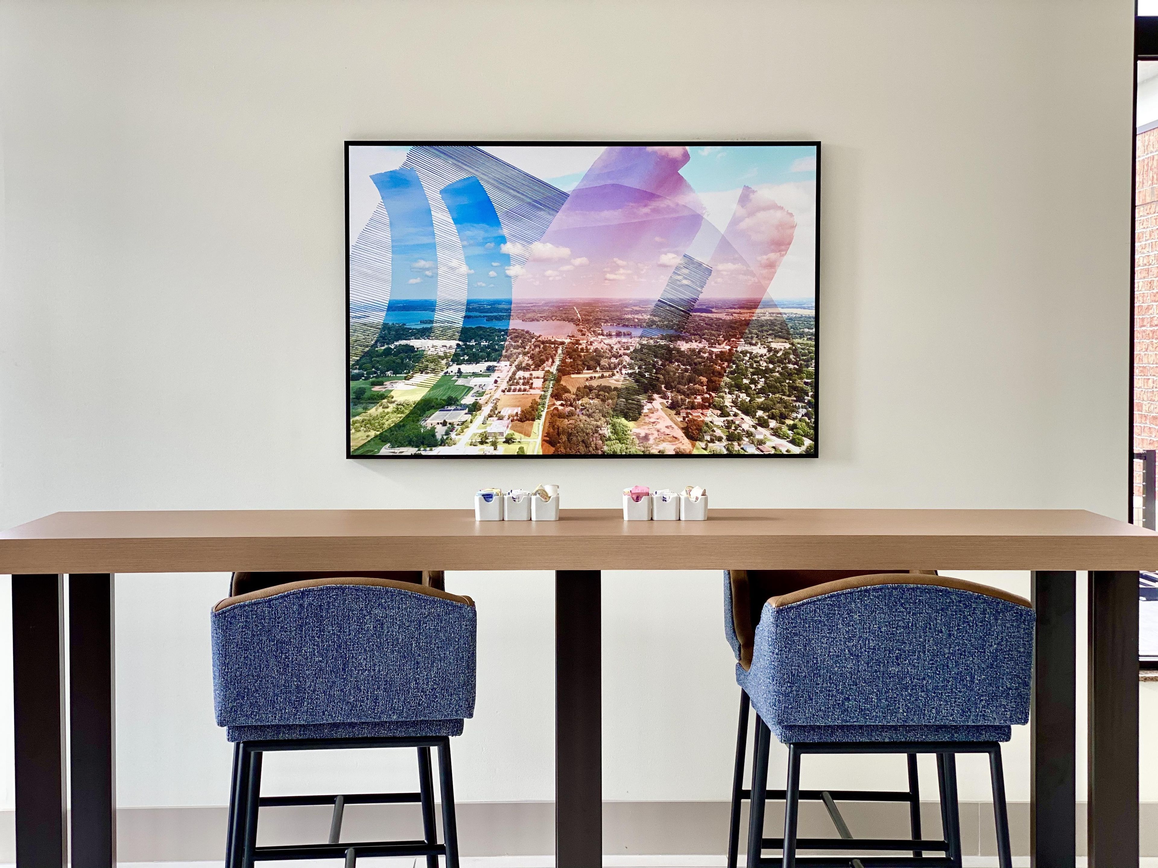 A framed aerial cityscape artwork on a wall above a wooden table with two blue chairs.