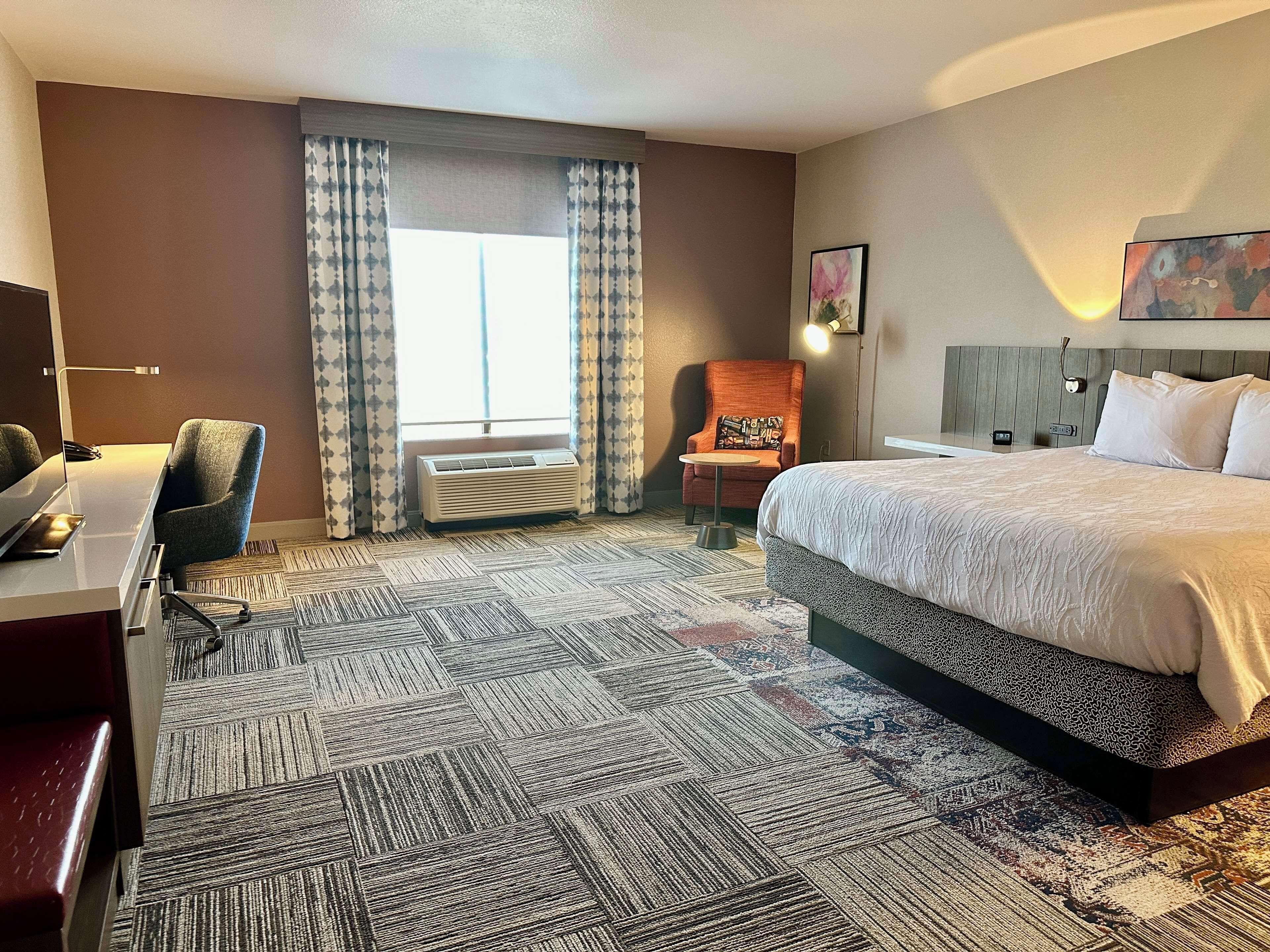 A hotel room with a bed, a desk, a chair, an air conditioning unit, and a window with curtains.