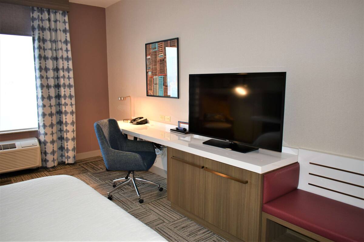 A hotel room with a bed, a desk with a computer and a TV, a chair, a red bench, and a window with curtains.