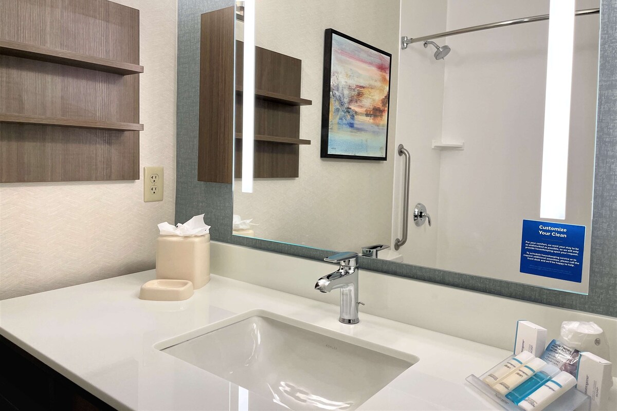 A modern bathroom with a large mirror, a sink, and a shower area. There is a sign on the wall and a framed picture above the mirror.