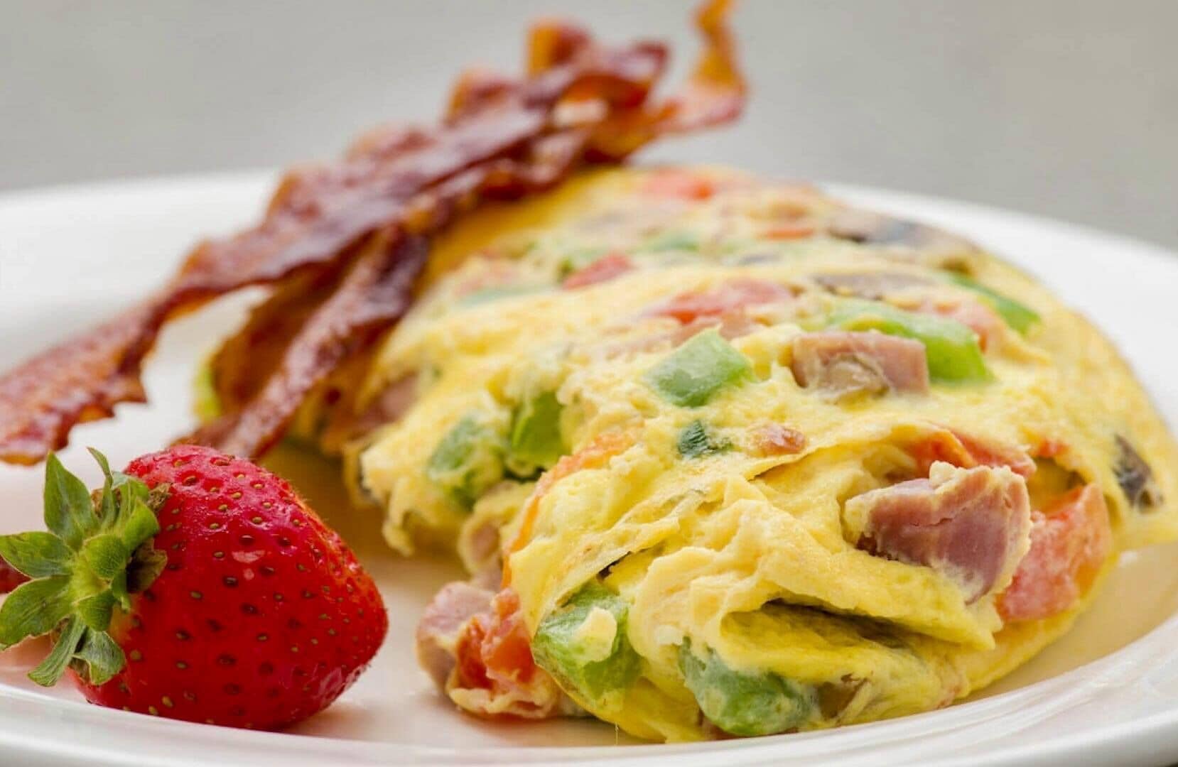 A plate with a breakfast omelette, bacon, and a strawberry.