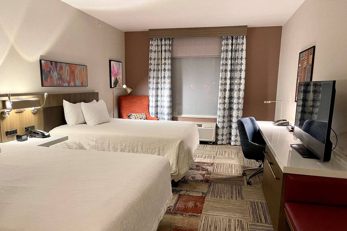 A hotel room with two beds, a desk, a chair, a TV, and a window with curtains.