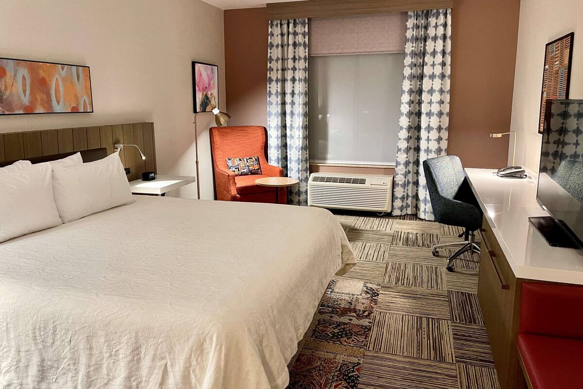 A hotel room with a large bed, a desk with a chair, a television, and a window with curtains.