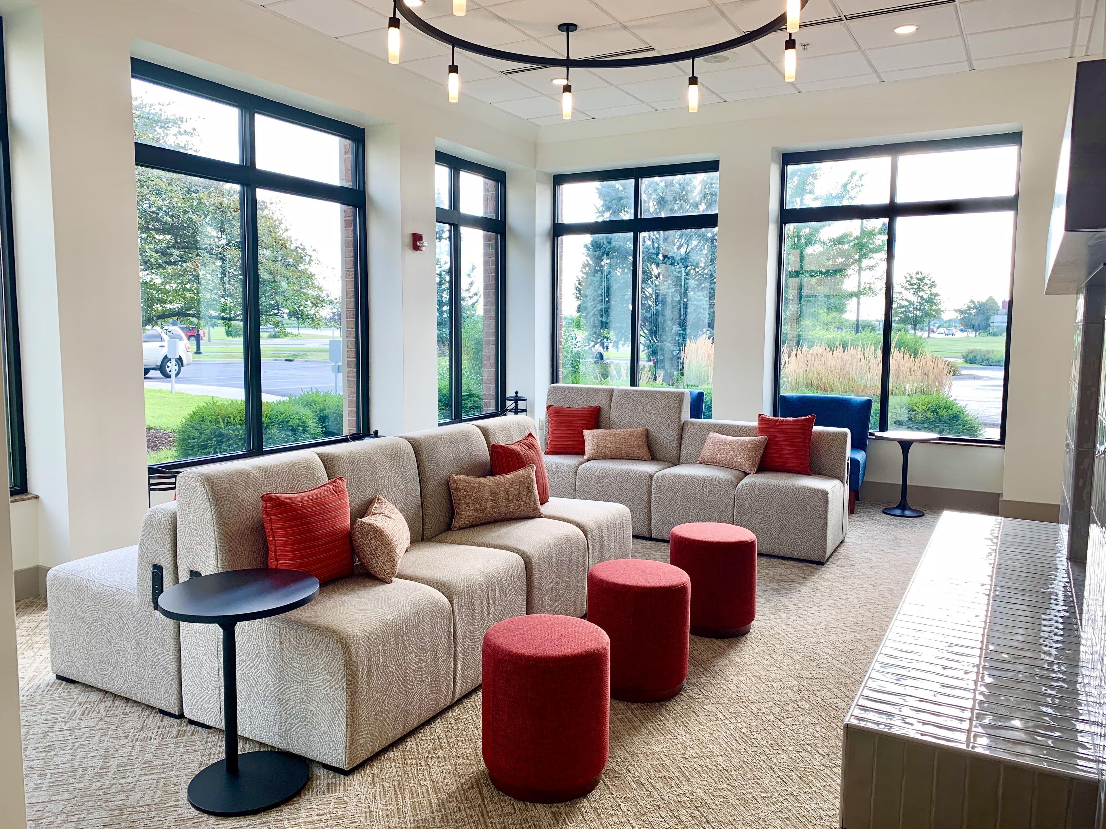 A spacious room with large windows, a sectional sofa, red ottomans, and a small round table.