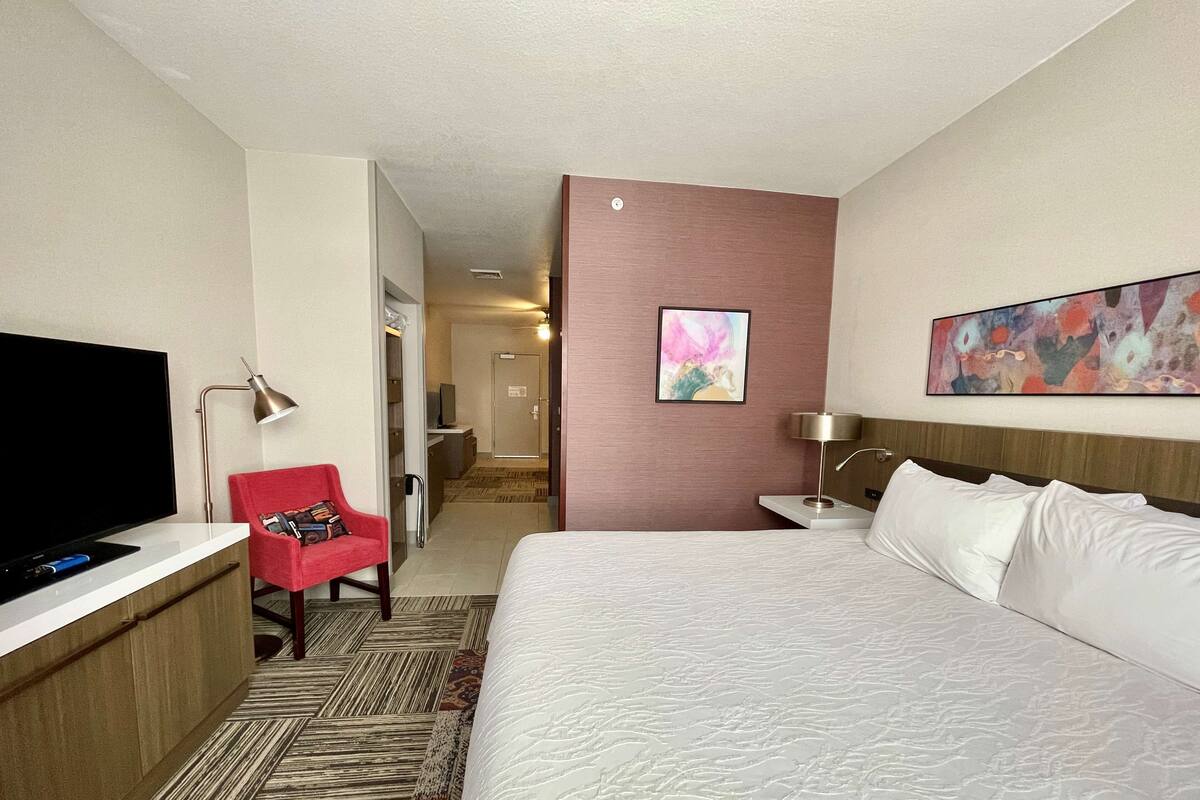 A hotel room with a large bed, a TV, a desk with a lamp, a red chair, and a painting on the wall.