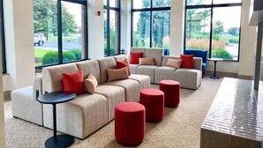 A spacious lobby with large windows, a sectional sofa, red ottomans, and a small table. - Hilton Garden Inn Oconomowoc (Oconomowoc)