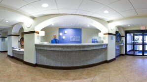 A reception area with a curved reception desk, a blue wall with a logo, and a person standing behind the desk. - Holiday Inn Express & Suites - Greenwood by IHG (Greenwood)