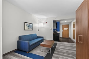 A modern hotel room with a blue sofa, a wooden coffee table, and a kitchenette in the background.