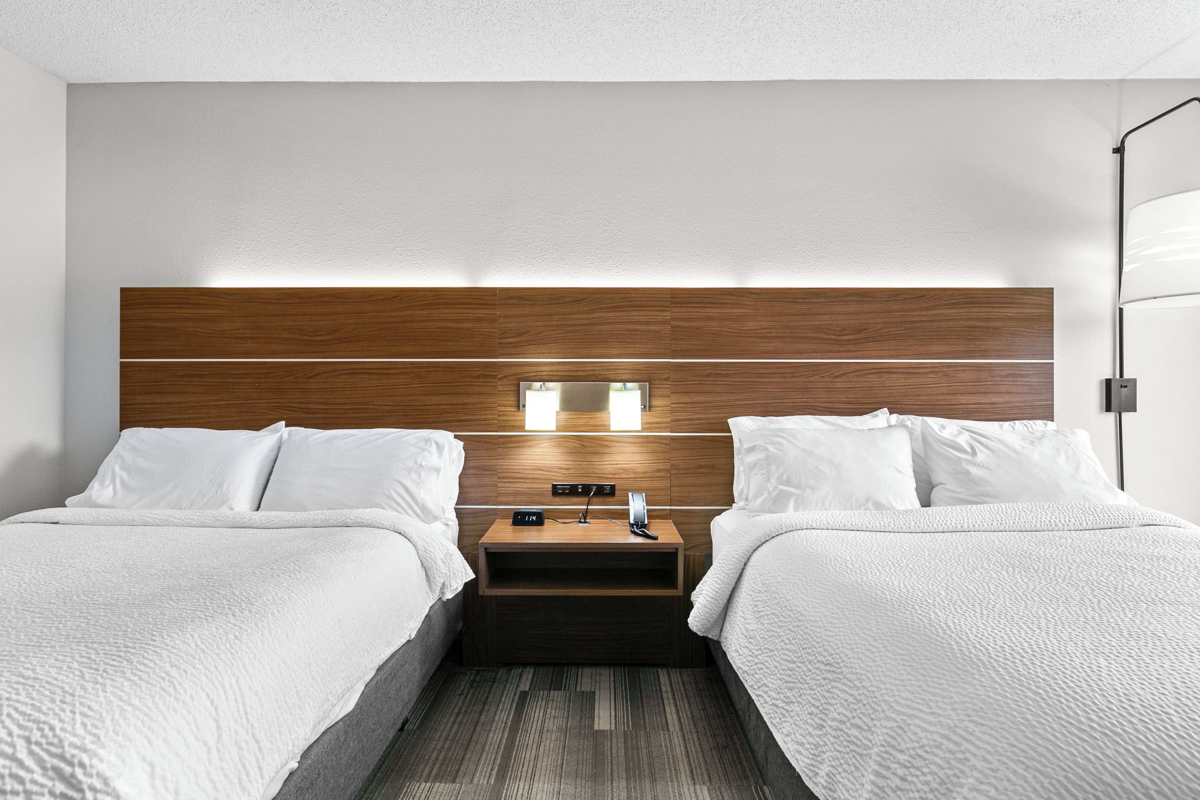 A hotel room with a wooden headboard, two beds with white bedding, a nightstand with a clock, and a bedside table.