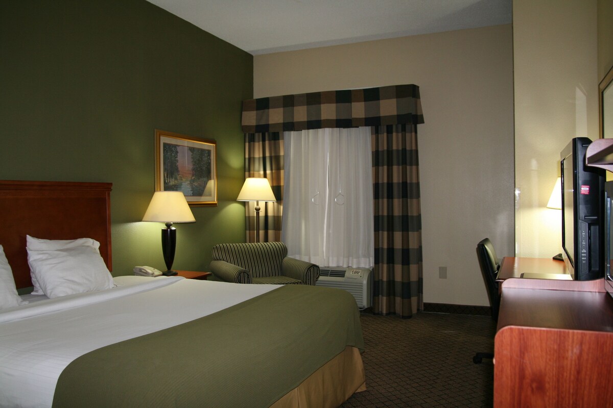 A hotel room with a large bed, two bedside lamps, a chair, a desk with a computer, and a window with curtains.