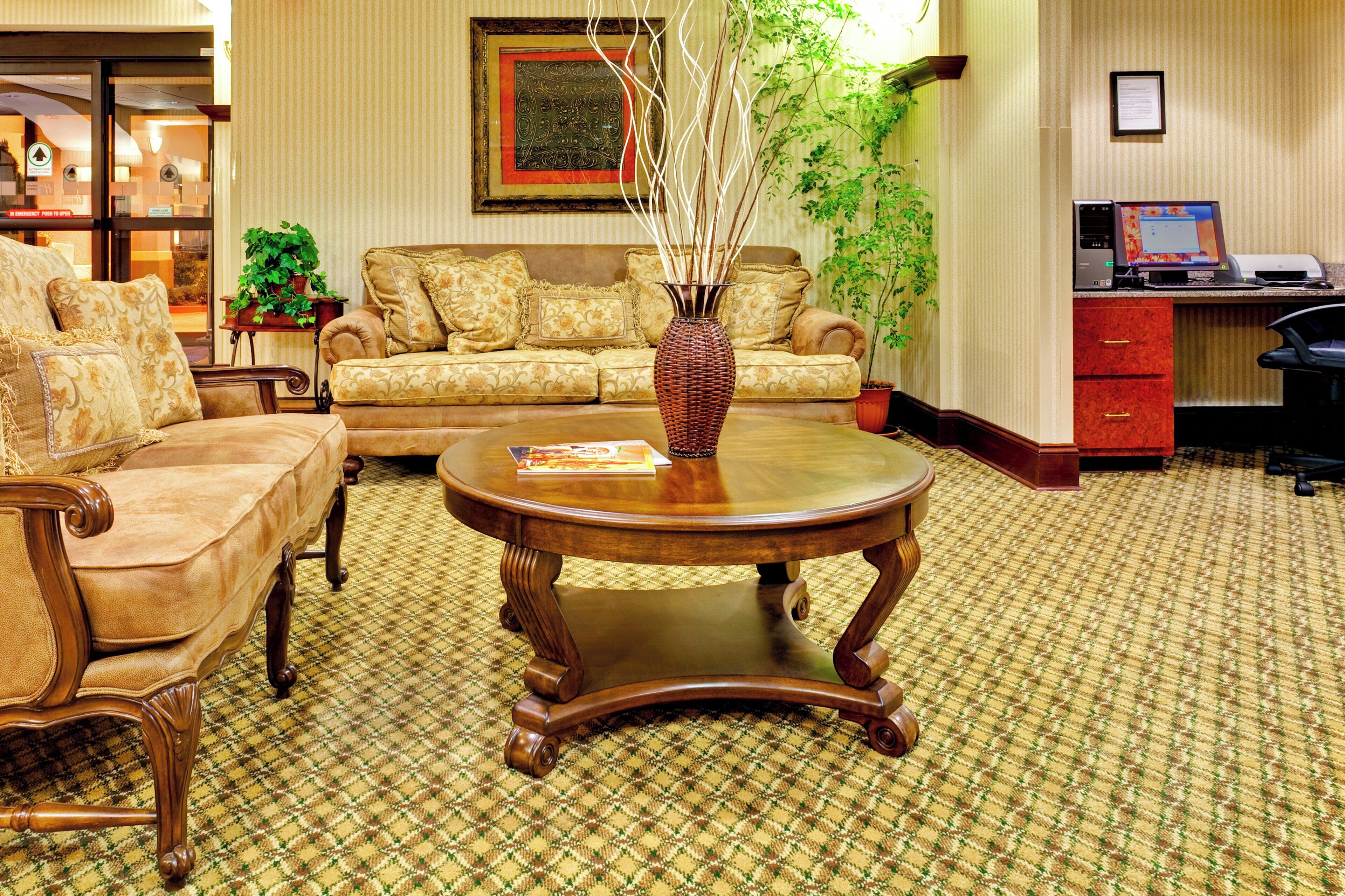 A hotel lobby with a patterned carpet, a round wooden coffee table, a sofa with floral cushions, and a potted plant.