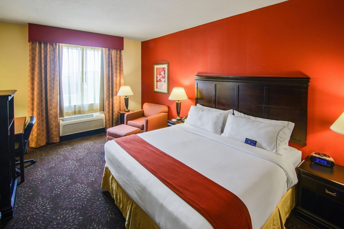 A hotel room with a large bed, a desk, a chair, a nightstand with a clock, and a window with curtains.
