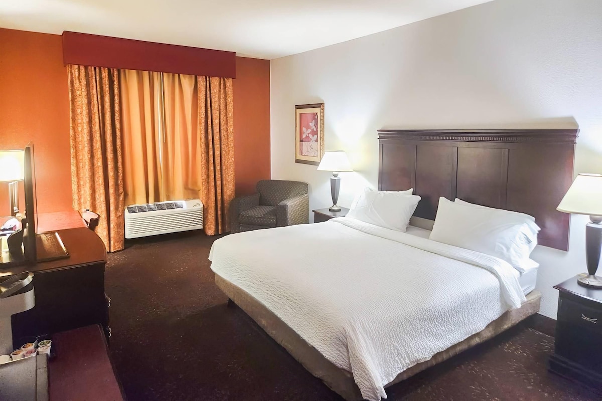 A hotel room with a large bed, two bedside lamps, a chair, a desk with a lamp, and a wall-mounted air conditioner.