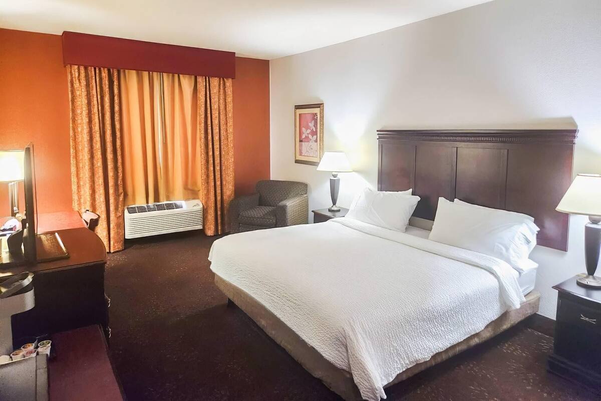 A hotel room with a bed, a chair, a desk with a lamp, a picture on the wall, and a window with curtains.