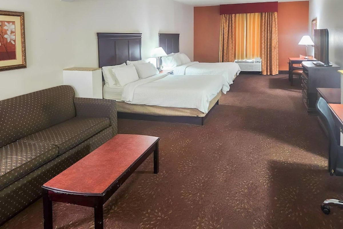 A hotel room with a sofa, a small table, a bed with two pillows, a desk with a lamp, and a television.