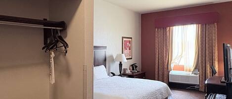 Une chambre d’hôtel comprenant un lit, un bureau avec un ordinateur, une chaise, un porte-manteau et une fenêtre avec des rideaux.