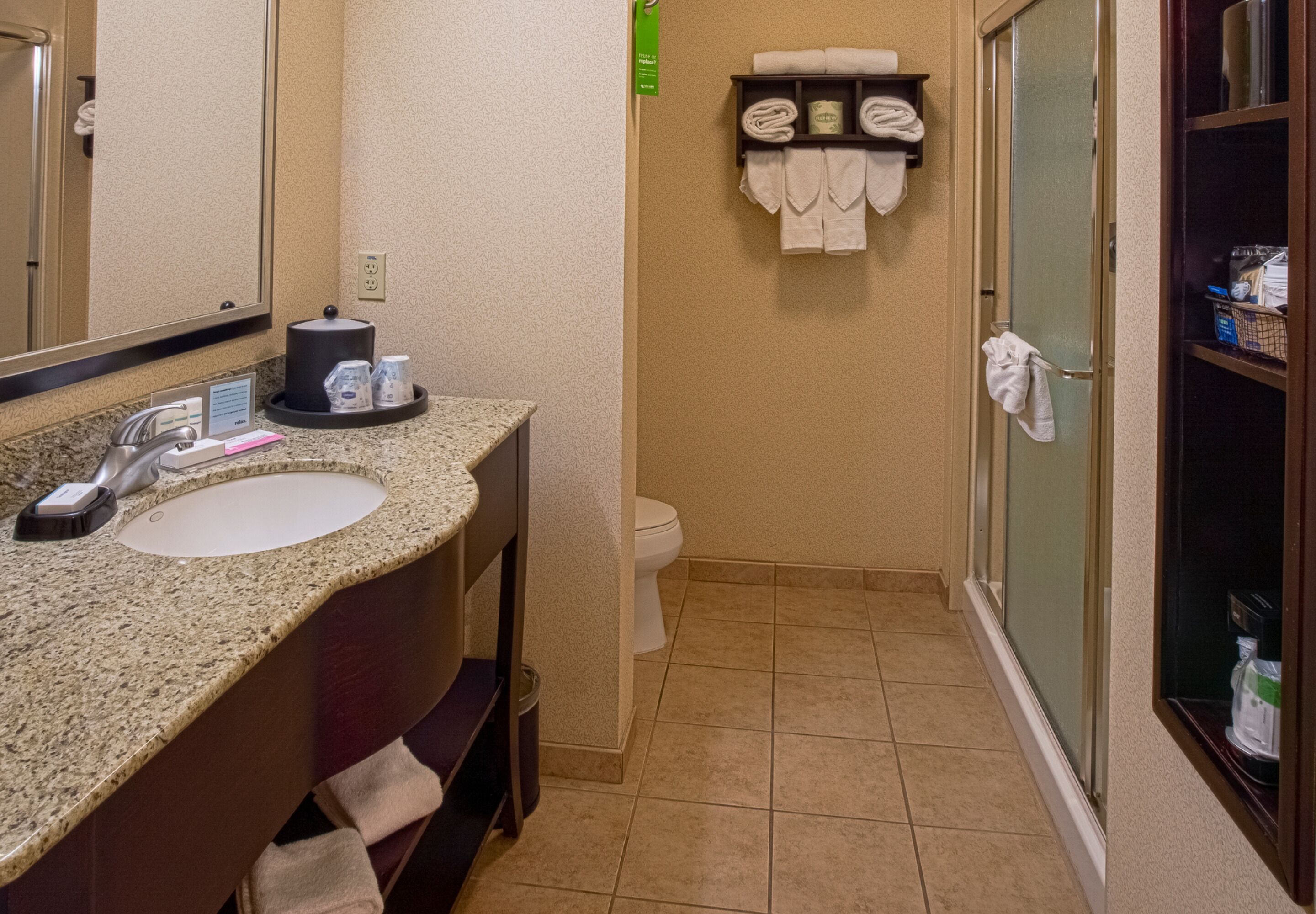 A bathroom with a granite countertop sink, a mirror, a towel rack with towels, a toilet, and a glass shower enclosure.