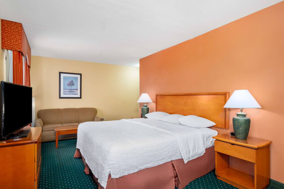 Une chambre d’hôtel comprenant un lit, une télévision, un canapé, deux tables de chevet avec des lampes et un tableau encadré au mur.