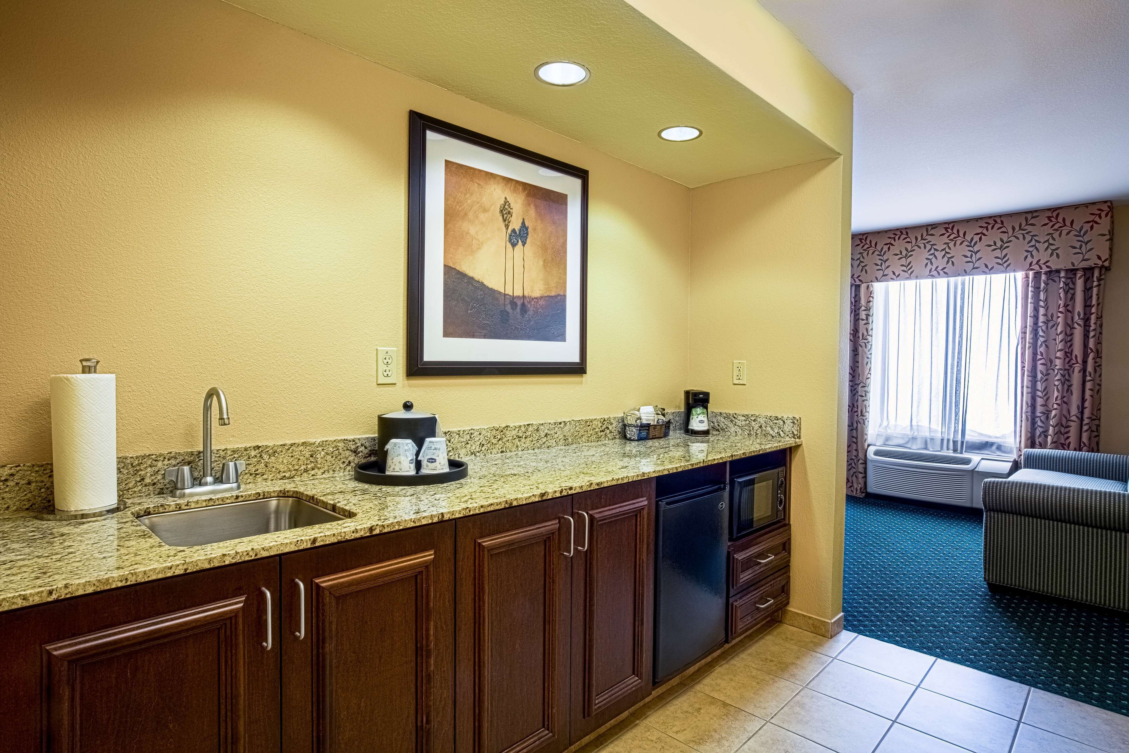 A hotel kitchen with a granite countertop, stainless steel sink, and wooden cabinets. There is a framed artwork above the sink and a window with curtains to the right.