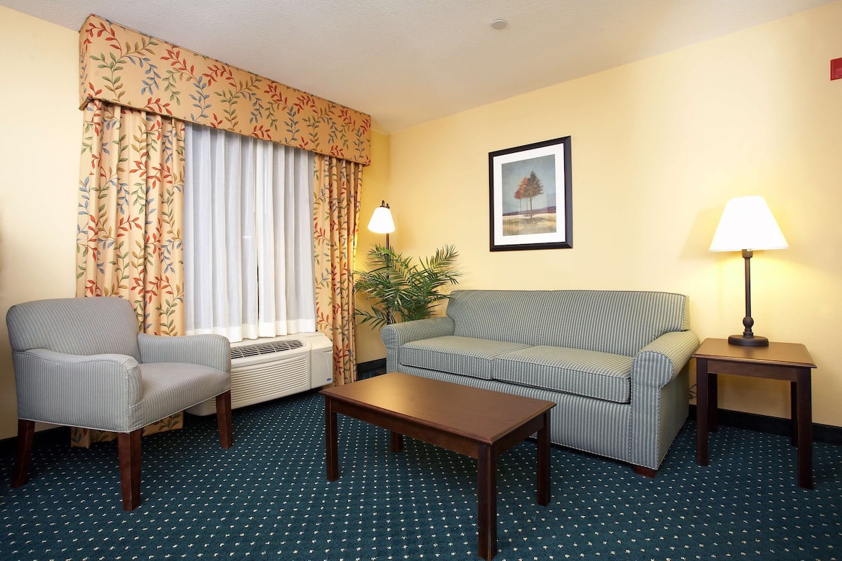 Une chambre d’hôtel avec un fauteuil à rayures, un canapé, une table basse en bois, un lampadaire et une grande fenêtre avec des rideaux.