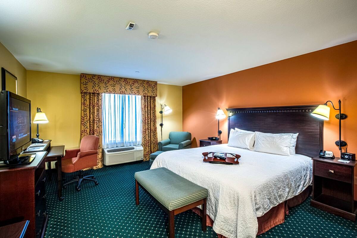 Une chambre d’hôtel avec un grand lit, un bureau avec un ordinateur, une télévision, deux chaises, un banc et une fenêtre avec des rideaux.