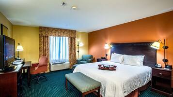 A hotel room with a large bed, a desk with a computer, a TV, two chairs, a bench, and a window with curtains.