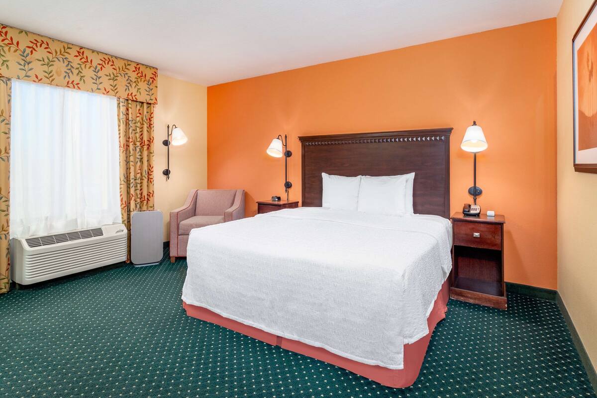 Une chambre d’hôtel avec un grand lit, deux lampes de chevet, une chaise, un radiateur et une fenêtre avec des rideaux.