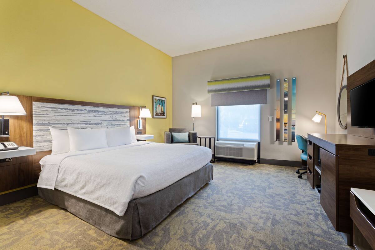 Une chambre d’hôtel avec un grand lit, des tables de chevet, un bureau, une chaise, une télévision et une fenêtre avec des stores.