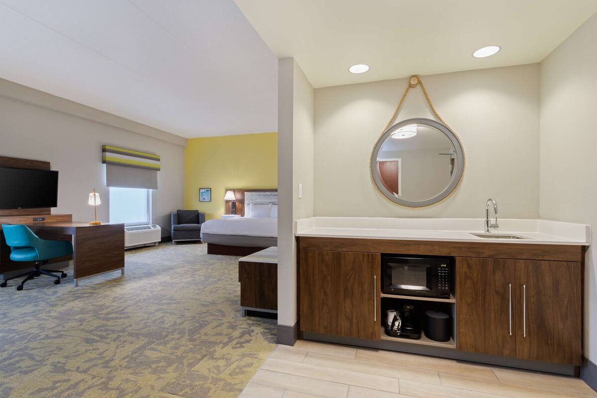 Une chambre d’hôtel avec un grand lit, un bureau avec une chaise, une télévision, une salle de bain avec un lavabo et un miroir, et un sol recouvert de moquette.