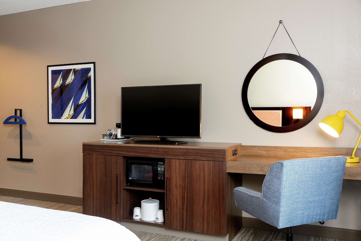 A hotel room with a bed, a TV on a wooden cabinet, a microwave, a blue chair, a round mirror, and a framed picture on the wall.