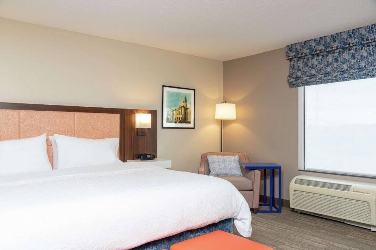 A hotel room with a bed, a desk, a chair, a lamp, a picture on the wall, and an air conditioning unit.