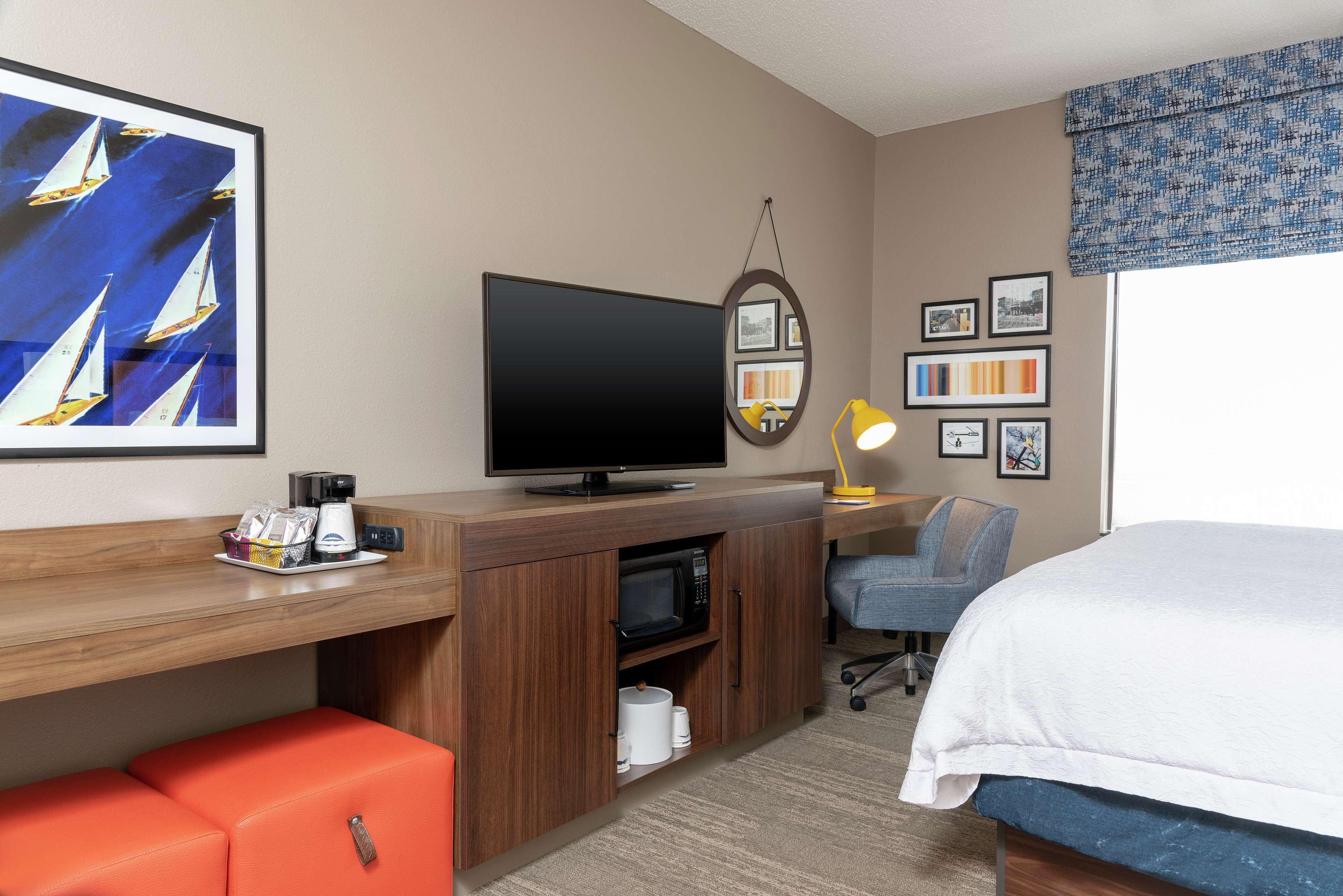 A hotel room with a bed, a desk with a flat-screen TV, a microwave, a small orange seating area, and a window with a view.