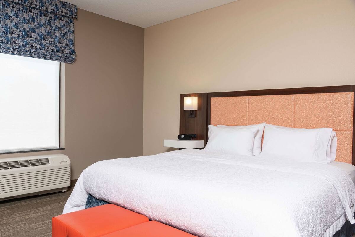 A hotel room with a bed, a bedside table with a lamp, a window with a view, and an air conditioning unit.