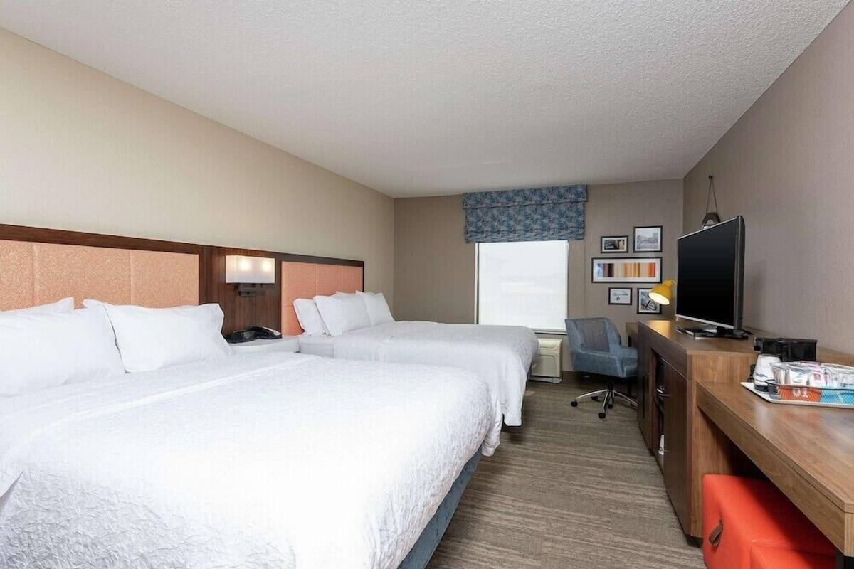 A hotel room with two beds, a desk, a TV, and a window with blinds.