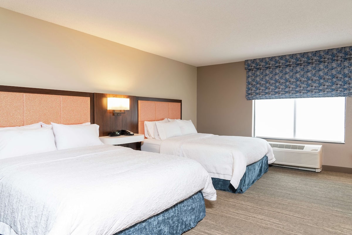 A hotel room with two beds, a nightstand, a lamp, and a large window with a patterned valance.