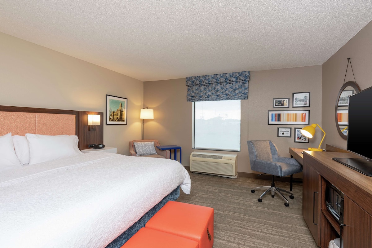 A hotel room with a large bed, a desk with a chair, a TV, and a window with a view.
