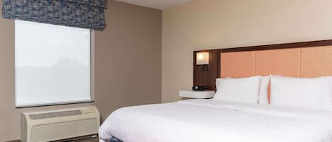 A hotel room with a large bed, an air conditioner, a nightstand with a lamp, and a window with a view.