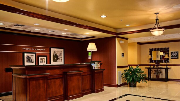 A reception area with a wooden reception desk, framed pictures, a lamp, and a potted plant. - Hampton Inn Enterprise (Enterprise)